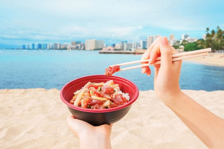 A lady sitting on a white sand beach eating a poke bowl in Hawaii