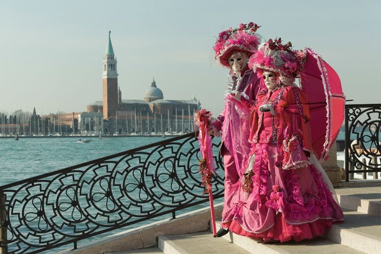 Two people in flamboyant pink costumes and masks on a bridge in Venice