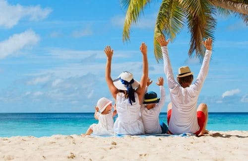 A family sits on a white sand beach with their hands in the air