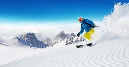 A person in yellow trousers and a blue jacket skiing down powdery snow-covered mountain side