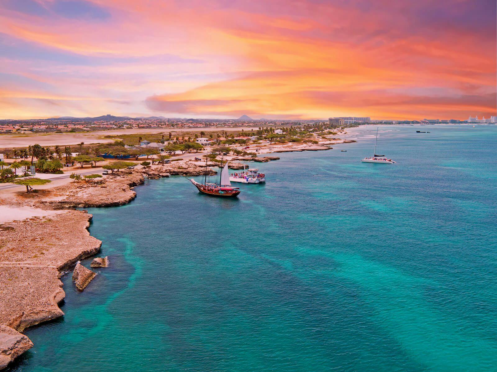Aruba coastline at sunset with sailboats in the water moored off of a rocky beach, with pink and orange clouds in the sky