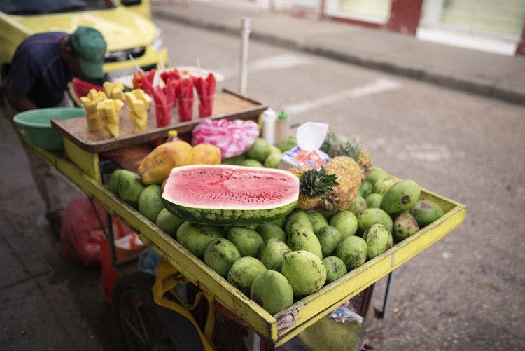 A man working on a fruit stall with watermelons, coconuts, and cups of melon and pineapple