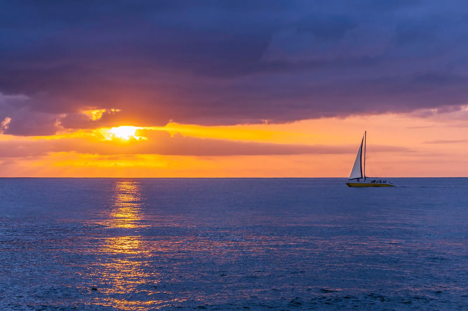 The sun sets over the Caribbean Sea in Barbados as a single sail boat drifts along 