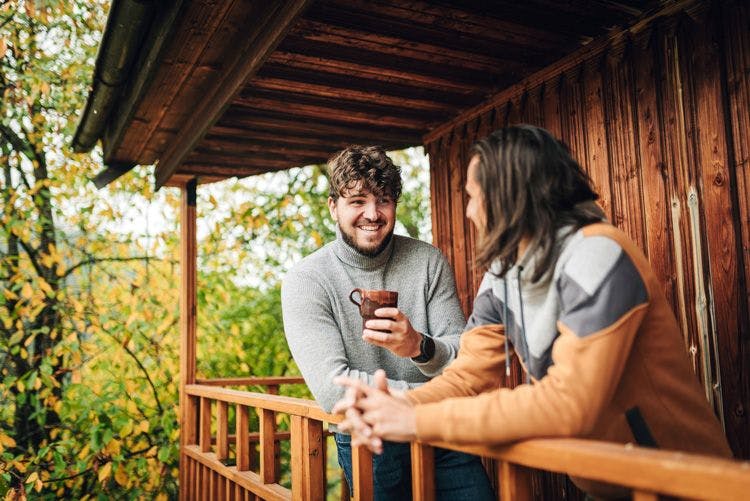 A young couple standing on a balcony of a cabin in the woods drinking coffee
