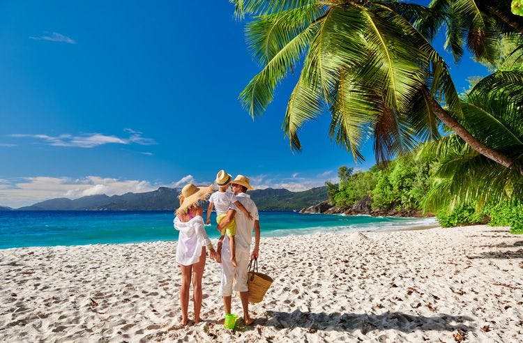 A family of three dressed in white walk along a white sand beach by palm trees