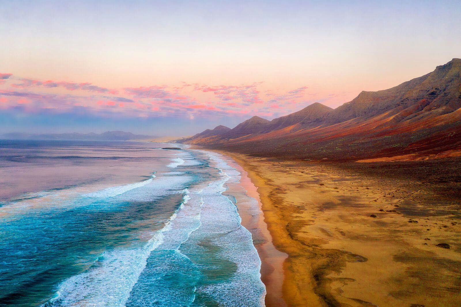 A golden sandy beach on Fuerteventura with dramatic volcanic mountains leading down into the Atlantic Ocean