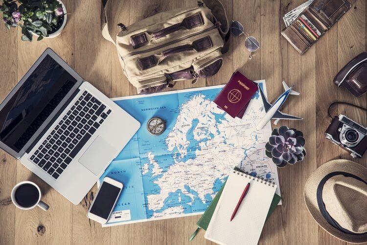 Travel planning on the floor with an open laptop, map of Europe, passport, notepad, satchel bag, camera, and model plane