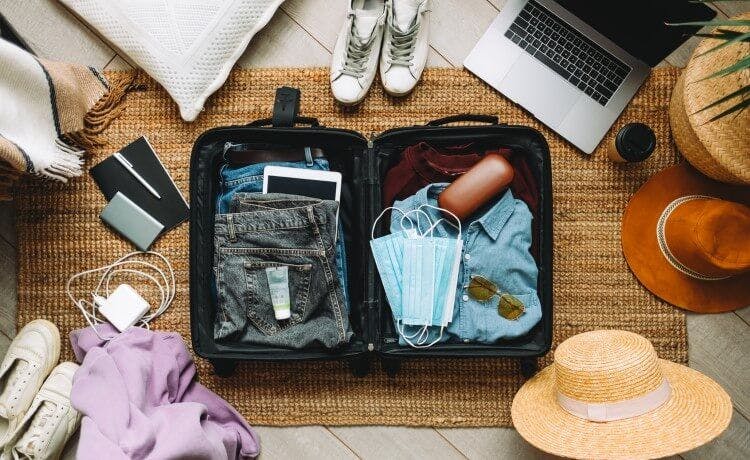 A suitcase, open, with vacation items including a hat, chargers, and shoes paced in and around the bag