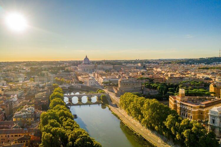 An aerial shot of the center of Roma, with a river flowing past historic buildings and tree-lined avenues
