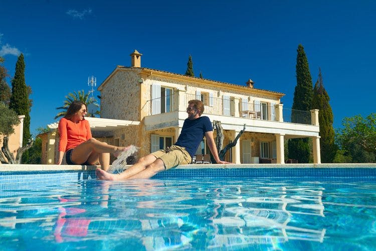 A couple sits on the edge of a private pool splashing the water with their feet in front of a villa
