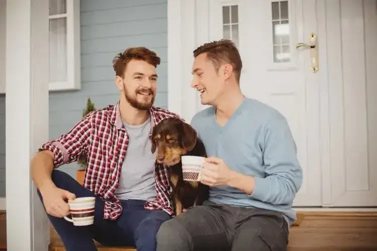 Two men sitting on a porch drinking coffee with a puppy
