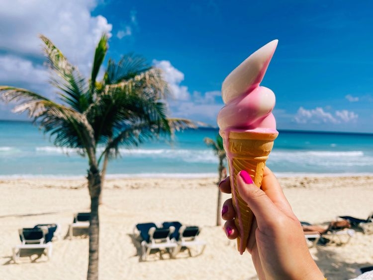 A woman holds and pink and white swirled ice cream cone in front of a white sand beach with a palm tree