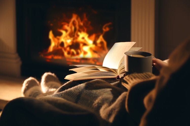 A woman sitting in a chair with a cup of tea and a book in front of a roaring fireplace