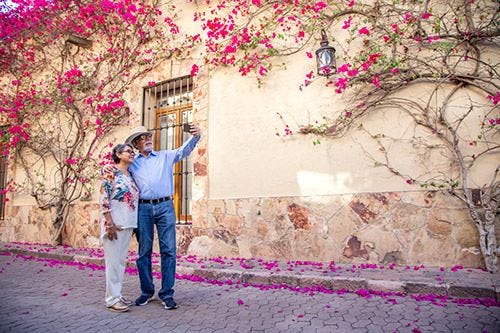 An older couple taking a photograph in front of a flower-covered wall in Europe