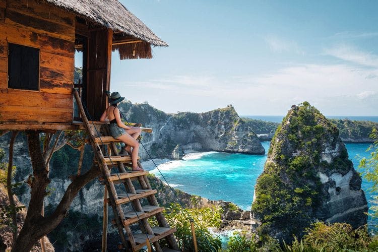 A woman siits on the steps of a wooden treehouse looking over a beach