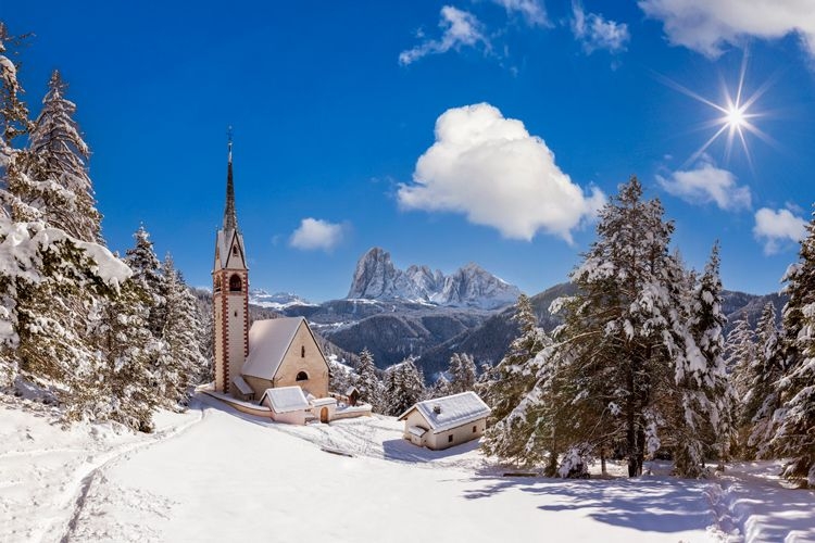 A small church in front of mountains in Italy with a thick blanket of snow on the ground