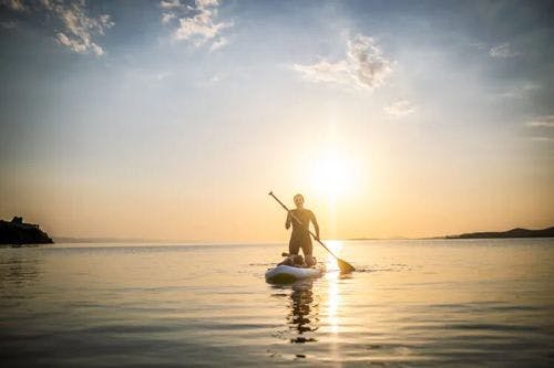 A person kneels on a paddleboard in the water at sunset