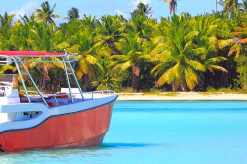A red and white fishing boat moored off of a white sand beach with a palm forest