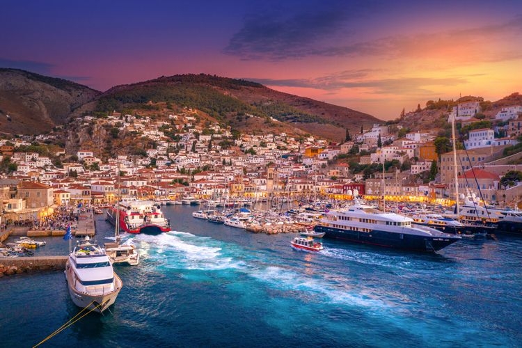 Sunset view of the small town of Hydra with ferries and fishing vessels in the harbor and small cube shaped buildings along the waterfront