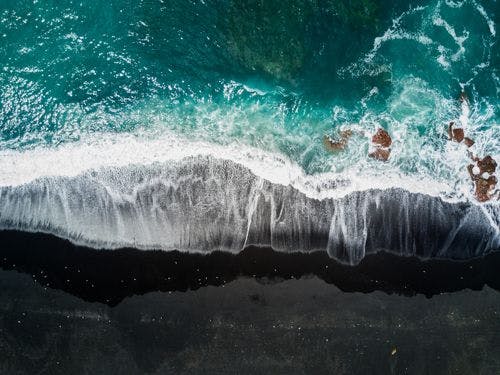 Aerial shot of a black sand beach with turquoise water and frothy surf