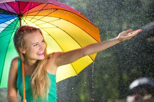 A woman standing under a rainbow umbrella laughing at the rain