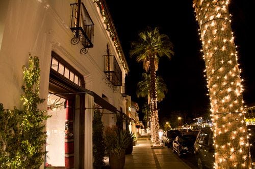 Boutique shops line a street at night with fairy lights wrapped around palm trees