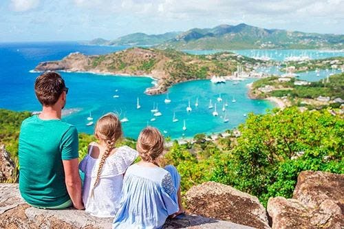 A family of a man and two young girsl sitting on a hill overlooking English Harbor in Antigua
