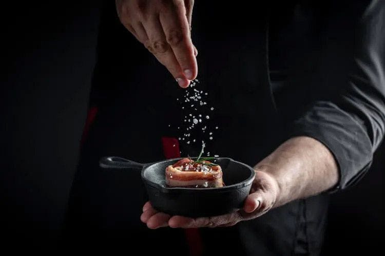 A chef holding a small dish of food and sprinkling salt on it