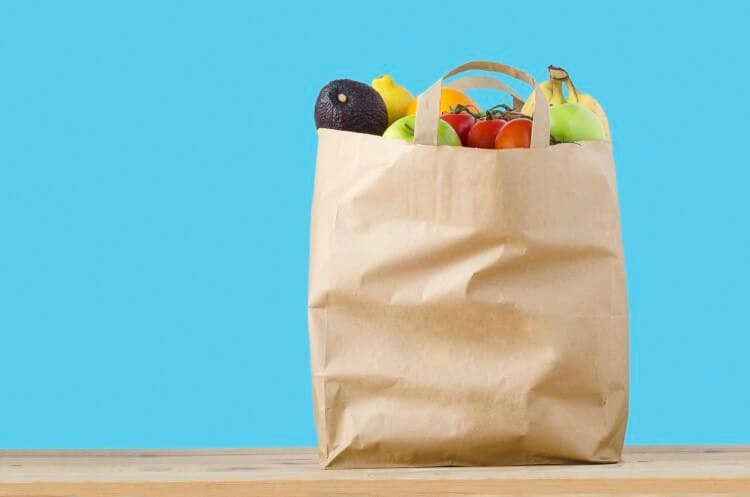 A brown paper bag filled with fruit and vegetables on a wooden table in front of a bright blue background