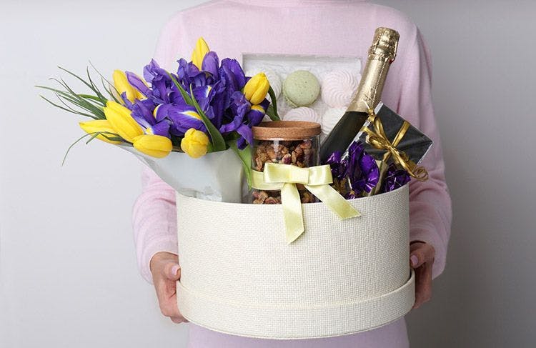 A person carrying a hamper box filled with gifts, including a bunch of flowers, champagne, chocolates, and more