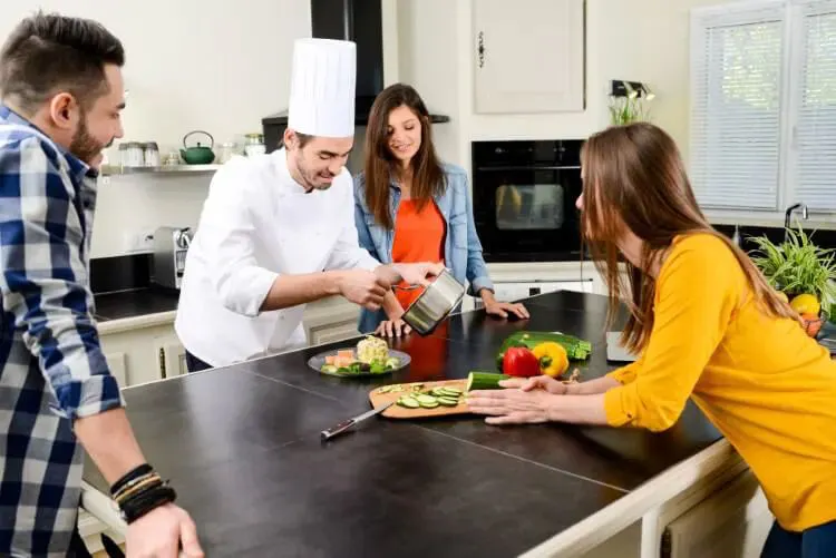 A chef prepares food for three people who are in the kitchen