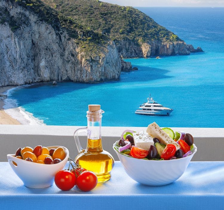 A bottle of olive oil, and bowls of olives and Greek salad on a table overlooking a Lefkada bay
