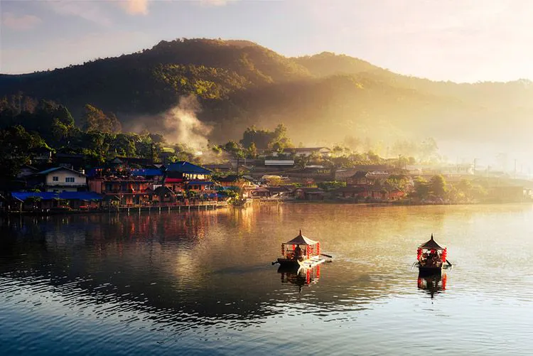 A misty morning on the Mekong River with two boats floating passed traditional Thai buildings
