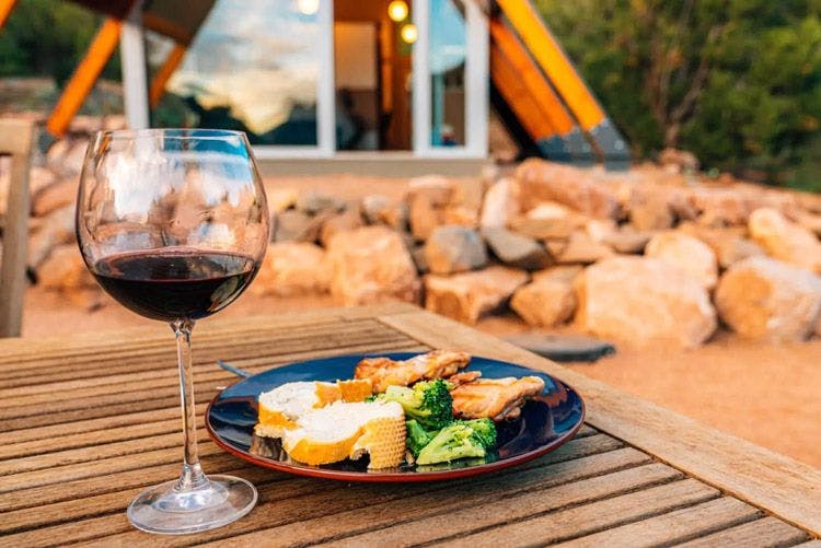 A plate of food and glass of wine on a wooden outdoor table by a cabin