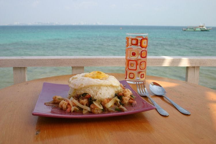 A plate of rice, vegetables and egg with a glass on a table by the sea
