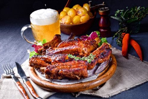 Smoked spare ribs served on a wooden plate with a glass of beer and a bowl of potatoes