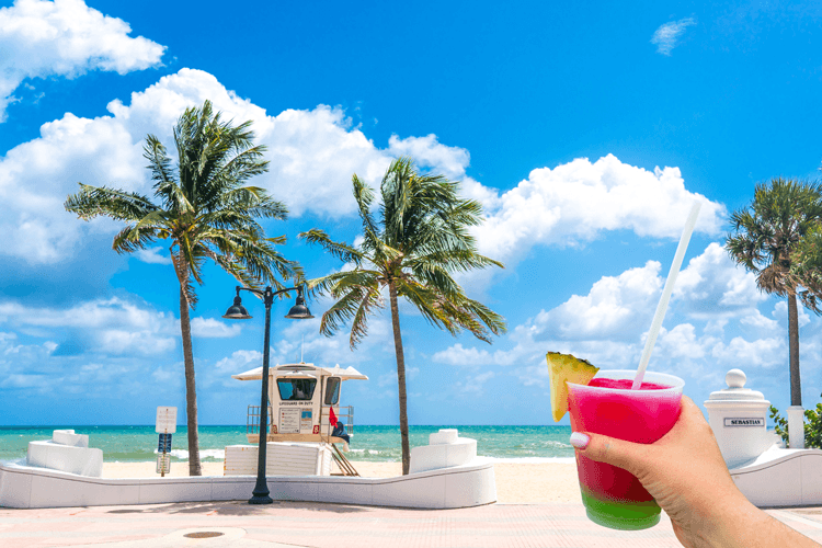 A person holding a frozen pink and green drink with a straw and pineapple wedge in front of a white sand beach with palm trees and lifeguard hut