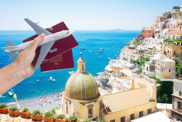 A hand holding two passports and a toy plane in front of an Amalfi Coast town