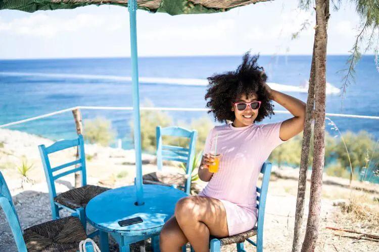 A woman sits at a table on the beach drinking an orange beverage