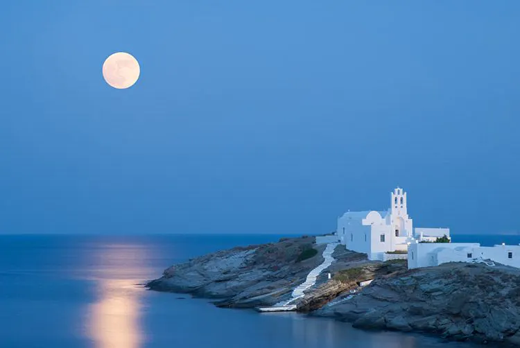 A full moon over a white monastery on the end of a spit of land jutting into the Aegean Sea