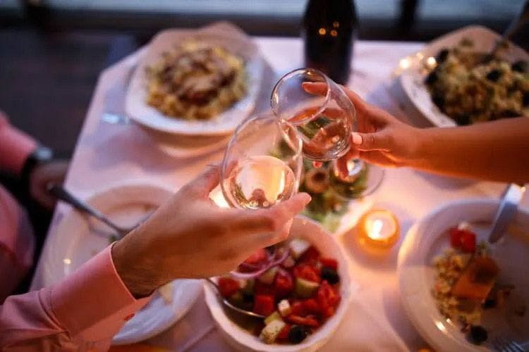 Two people sit at a table laid for dinner clinking wine glasses