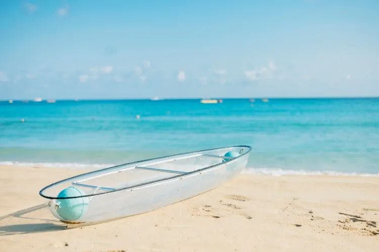 A clear kayak resting on white sand by turquoise sea