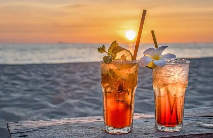 Two orange cocktails on a table by a white sand beach at sunset