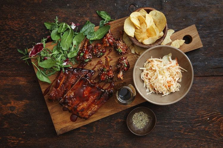 Overhead shot of barbecued meat on a wooden board with sides of coleslaw and chips