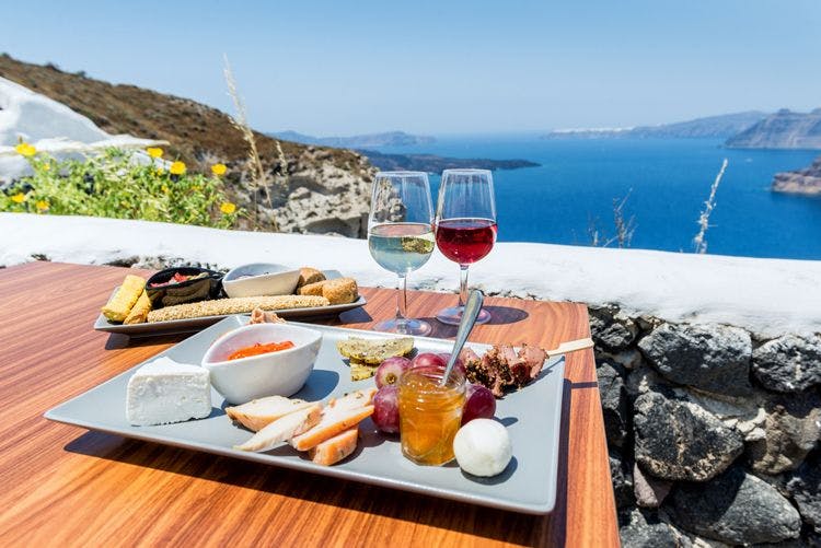 Plate of bread, fruit, honey, and cheese with two glasses of wine overlooking the sea in Santorini