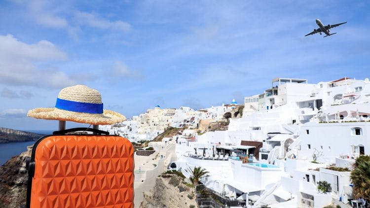 Santorini village with orange suitcase and sunhat in front and plane flying overhead