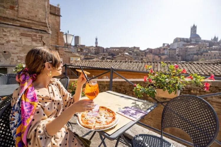 A woman sitting at a restaurant with a pizza and aperol spritz drink looking out over the city of Siena