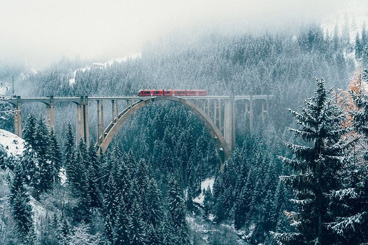 A red train going across a tall viaduct bridge in a Swiss forest in winter