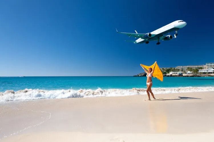 A woman holds a yellow towel above her head as she walks along a beach and a plane flies overhead