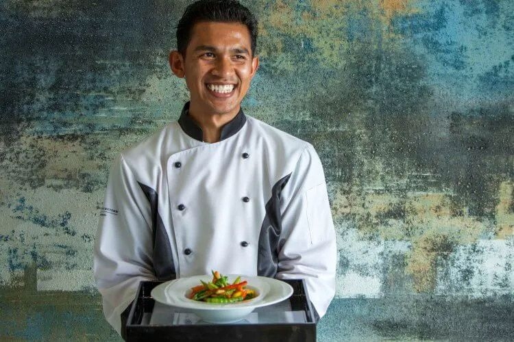 A chef wearing a white jacket smiles as he holds a plate of food